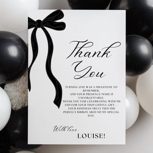 May include: A white card with a black ribbon and the words "Thank You" in elegant script. The card is surrounded by black and white balloons and decorative accents. The text on the card reads "Turning one was a milestone to remember..." and is signed "Louise!"