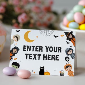 Może przedstawiać: Biała karta o tematyce Halloween z czarnym napisem "ENTER YOUR TEXT HERE". Karta zawiera ilustracje czarnego kota, dyni, wiedźmy i innych postaci z Halloween. Dwa cukierki znajdują się na pierwszym planie.