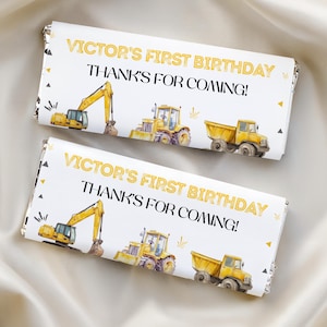 Könnte beinhalten: Zwei rechteckige Schokoriegelverpackungen mit weißem Hintergrund und gelben Baustellenfahrzeug-Illustrationen, darunter ein Bagger, ein Traktor und ein Muldenkipper. Der Text lautet VICTOR'S FIRST BIRTHDAY und THANK'S FOR COMING!