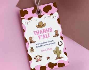 Könnte beinhalten: Ein rechteckiges Geschenkanhänger aus Papier mit einem rosa-braunen Kuhfleckenmuster am Rand und einem weißen Zentrum. Er zeigt Illustrationen von einem Cowboyhut, Stiefeln, einem Geldsack, einer Kuh, einem Kaktus und einem Hufeisen sowie den Text "THANKS Y'ALL FOR GALLOPING ON BY TO CELEBRATE MY FIRST RODEO Love, Alice."