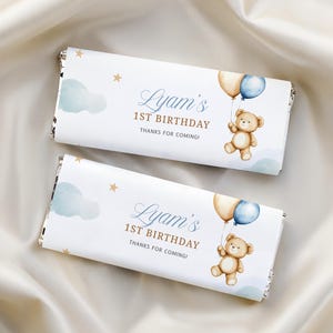 May include: Two rectangular chocolate bar wrappers featuring a watercolor illustration of a brown teddy bear holding a beige and blue balloon. The text reads Lyam's 1ST BIRTHDAY THANKS FOR COMING! with light blue clouds and gold stars on a white background.