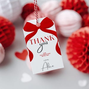 Puede incluir: Una etiqueta de regalo rectangular blanca con un gráfico de un lazo rojo grande en la parte superior. El texto dice THANK you FOR CELEBRATING WITH ME! LOVE, Alice. La etiqueta está suspendida por un cordel rojo y blanco a través de un orificio.