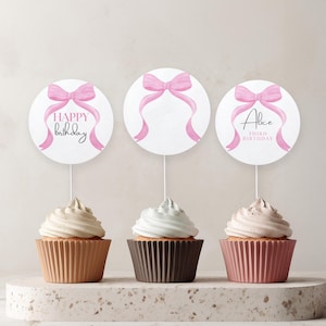 Op de afbeelding: Drie cupcakes met witte frosting en decoratieve toppers. De toppers zijn witte cirkels met roze strikken en linten. Eén zegt "Happy Birthday", een andere heeft een naam en "Third Birthday". De cupcakes zitten in bruine, donkerbruine en roze cupcakevormpjes.