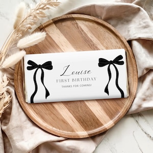 Op de afbeelding: Een witte chocoladereep met zwarte strikillustraties en de tekst "Louise EERSTE VERJAARDAG BEDANKT VOOR JE KOMST!" De reep ligt op een houten dienblad met een beige stoffen achtergrond.