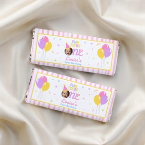 May include: Two rectangular chocolate bar wrappers featuring a white background with pink and white vertical stripes on the edges. The design includes colorful balloons, small stars, and the text "FUN TO BE one Louise's First Birthday" with a photo of a child.