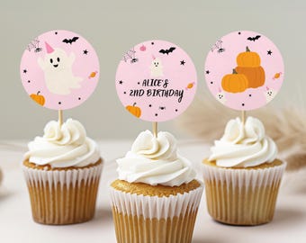 Op de afbeelding: Drie cupcakes met ronde roze papieren prikkers met Halloween-illustraties. De prikkers tonen een geest met een eenhoornhoorn, oranje pompoenen, zwarte vleermuizen, spinnen en sterren. Eén prikker bevat de tekst Alice's 2nd Birthday.