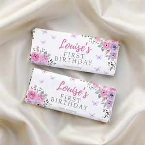 May include: Two rectangular candy bar wrappers featuring a white background with pink and purple floral arrangements, small butterflies, and the text "Louise's FIRST BIRTHDAY" in pink and gray script and block lettering, with "Thanks for coming!" below.