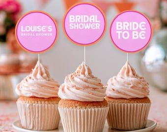 May include: Three cupcakes with light pink frosting in white paper liners, each topped with a round pink sign on a stick. The signs feature orange borders and white text reading "Louise's Bridal Shower," "Bridal Shower," and "Bride to Be."