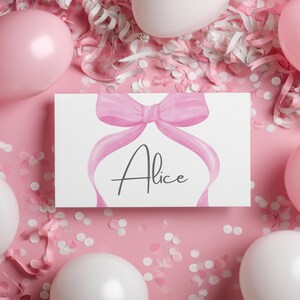 以下が含まれることがあります： ピンク色の水彩画のリボンと筆記体で「Alice」という名前が書かれた白いカード。カードはピンクと白の風船、紙吹雪、ストリーマーに囲まれ、お祝いの雰囲気を醸し出しています。