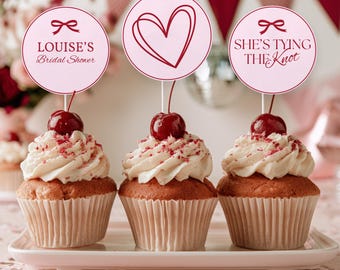 May include: Three cupcakes with white frosting, red sprinkles, and a cherry on top, each featuring a round paper topper. The toppers display a red bow with the text "LOUISE'S Bridal Shower", a red heart outline, and a red bow with "SHE'S TYING THE KNOT".