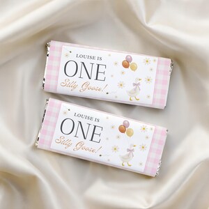 May include: Two rectangular chocolate bar wrappers featuring a pink and white gingham pattern on the edges. The central white label displays the text "LOUISE IS ONE Silly Goose!" in black and gold, accompanied by a goose illustration with balloons.