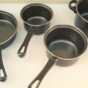 May include: A set of four black pots with black handles. The pots are of different sizes and are arranged on a white surface.