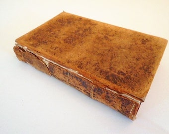 May include: A vintage book with a worn, textured leather cover. The cover is a warm brown with darker brown spots and signs of age. The spine shows visible stitching and wear, suggesting it is an antique or well-loved item.