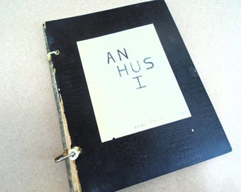 May include: A black, textured binder with a white rectangular label on the front cover. The label features the black printed text AN HUS I and the handwritten name Lewis Smith. The binder is secured with a metal ring fastener on the left side.