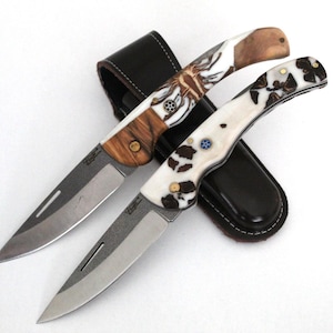 Könnte beinhalten: Zwei Klappmesser mit gemusterten Griffen, eines mit einem braunen und weißen Muster und das andere mit einem weißen und schwarzen Muster. Die Messer befinden sich in einer schwarzen Lederscheide.