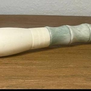 May include: A calligraphy brush with a light brown bristle head and a white handle. The handle transitions to a light green and gray ridged section, and ends with a white tip. The brush is lying on a wooden surface.
