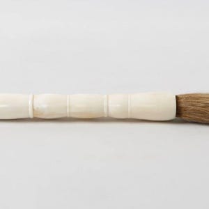 May include: A calligraphy brush with a cream-colored handle and a brown brush head. The handle is segmented with a small black string attached to the end. The brush is designed for detailed writing and artistic expression.