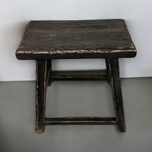 Puede incluir: Un taburete de madera rústico con un acabado pintado de negro y un aspecto desgastado. El taburete tiene un diseño sencillo con una parte superior rectangular y cuatro patas.