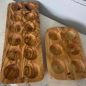 May include: Two wooden serving trays with carved circular indentations. The trays are made of olive wood, displaying a natural wood grain pattern with varying shades of brown and tan. One tray is rectangular with 12 indentations, and the other is square with 6.