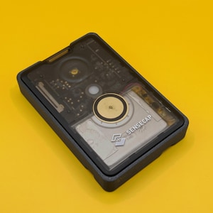 May include: A black plastic case with a clear top, revealing a circuit board with a gold-colored sensor and the text "SENSECAP" printed on the board.