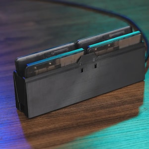 May include: A black electronic device with two slots, each holding a rectangular card. The device has a sleek, modern design with a matte finish. A black cable extends from the back. The device sits on a wooden surface.