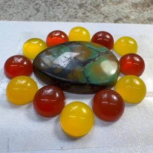 May include: A large, oval-shaped turquoise cabochon with dark brown matrix veins, surrounded by a circular arrangement of twelve smaller, smooth, dome-shaped cabochons in alternating shades of bright yellow and deep orange-red.