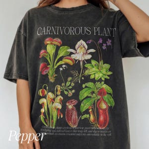 May include: Dark gray t-shirt featuring a botanical illustration of various carnivorous plants. The shirt includes the text "CARNIVOROUS PLANT" at the top and a description of the plants at the bottom. The design showcases detailed drawings of Venus flytraps and pitcher plants.