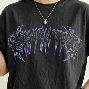 May include: A dark grey t-shirt featuring the text "CUSTOM TEXT" in a dripping, stylized font with purple and green highlights. A gold necklace with a heart-shaped pendant is visible. The shirt has a washed-out look.