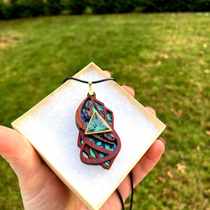Könnte beinhalten: Eine Holz-Halskette mit einem dreieckigen grünen Edelstein, der von kleineren blauen Edelsteinen umgeben ist. Der Anhänger ist aus Holz mit einer dunkelbraunen Oberfläche und hat eine goldfarbene Metallöse.