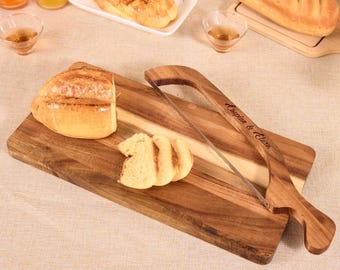 May include: A wooden bread cutting board with a loaf of bread, sliced bread, and a bread saw. The bread saw has a wooden handle with the text "Caspian & Clara". The board is made of a light brown wood.