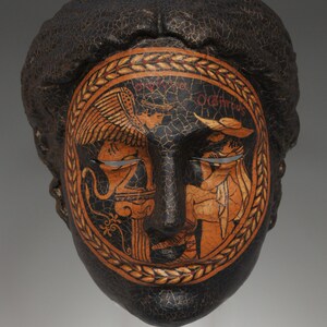 May include: A black mask with a cracked texture and a painted design of a face surrounded by a wreath. The face has closed eyes and a wreath of leaves around it. The text "Orgitus" is visible on the wreath.