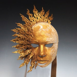 May include: A gold and white masquerade mask with a detailed design of flames or sun rays. The mask is on a stand and has a textured surface.