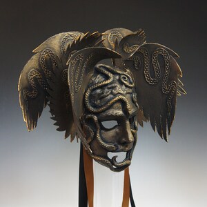 May include: A gold-colored mask with a snake design and wings. The mask has a human face with a long nose and a wide mouth. The wings are made of leather and have a textured surface.