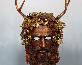 May include: A full-face mask featuring a textured, cracked brown finish depicting a bearded face. The mask is adorned with metallic gold oak leaves, acorns, and mistletoe sprigs, and is topped with two prominent, branching deer antlers.