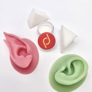Pode incluir: Um modelo de orelha de silicone rosa e um modelo de orelha de silicone verde, duas esponjas de maquilhagem triangulares brancas e um pequeno recipiente circular com tampa vermelha, logótipo dourado e pó branco no interior.