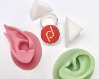 May include: A pink silicone ear model and a green silicone ear model, two white triangular makeup sponges, and a small circular container with a red lid featuring a gold logo and white powder inside, arranged on a plain white surface.