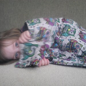 May include: A young person with long brown hair is lying on a light gray surface. They are wearing a colorful blanket with a pattern of owls and stars. The blanket is made of soft fabric and has a white background.