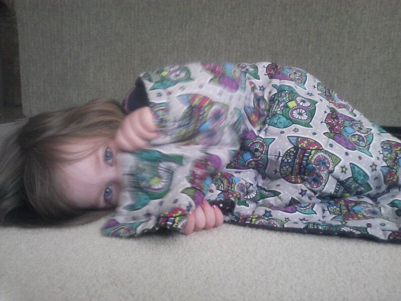 May include: A young person with long brown hair is lying on a light gray surface. They are wearing a colorful blanket with a pattern of owls and stars. The blanket is made of soft fabric and has a white background.