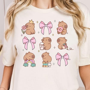 May include: A white t-shirt with a graphic print of nine adorable capybaras wearing bows and engaging in various activities, such as drinking a milkshake, reading a book, and holding a donut.