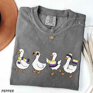May include: Pepper-colored t-shirt featuring a line of cartoon ducks with non-binary pride accessories. The ducks wear scarves, sunglasses, and hats in the colors of the non-binary flag. A silver necklace with a pendant is also visible.