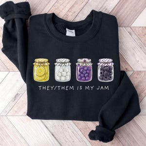 May include: Black sweatshirt featuring four illustrated jam jars filled with lemons, sugar cubes, purple grapes, and blackberries. The text "THEY/THEM IS MY JAM" is printed below the jars. The sweatshirt has a crew neck and long sleeves.