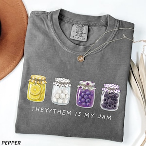 May include: Pepper-colored t-shirt with a graphic of four jam jars, each filled with different items: lemon slices, sugar cubes, blueberries, and blackberries. The text "THEY/THEM IS MY JAM" is printed below the jars. A silver necklace with a pendant is draped on the shirt.