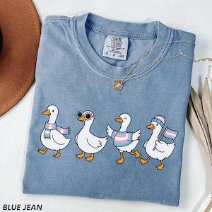 May include: A blue jean Comfort Colors t-shirt featuring four cartoon ducks, each with unique accessories and a transgender pride theme. The ducks are wearing sunglasses, scarves, and holding a flag. A gold necklace is draped across the shirt.