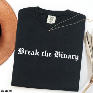 May include: Black t-shirt with the phrase "Break the Binary" in white gothic lettering. A gold necklace with a star pendant is draped across the shirt. A brown hat is visible in the upper left corner.