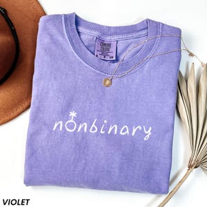 May include: A violet Comfort Colors t-shirt with the word "nonbinary" in white script. A gold necklace with a pendant rests on the shirt. A brown hat and dried plant are in the background.