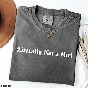 May include: A gray Comfort Colors t-shirt with the text "Literally Not a Girl" in white gothic lettering. A silver necklace with a round pendant rests on the shirt. A brown hat is partially visible.