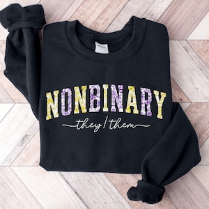 May include: Black sweatshirt with the word "NONBINARY" in yellow and purple floral letters. Below the word is the phrase "they/them" in a cursive font. The sweatshirt has a crew neck and long sleeves.