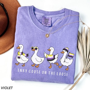 May include: Violet-colored t-shirt with the text "ENBY GOOSE ON THE LOOSE." The shirt features a graphic of four cartoon geese, each with non-binary pride accessories. A gold necklace is draped on the shirt.