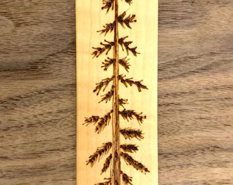 Könnte beinhalten: Ein Holz-Lesezeichen mit einem eingebrannten Design eines Kiefernbaums. Das Lesezeichen ist hellbraun und hat oben ein Loch zum Aufhängen.