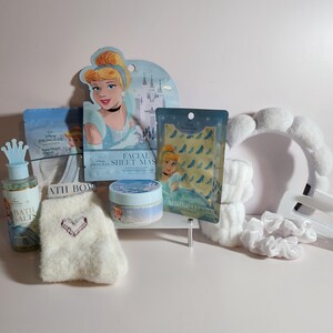 May include: A Disney Princess themed self-care set including a bottle of bath salts with a crown cap, a Cinderella facial sheet mask, a Cinderella under-eye patch packet, a jar of body scrub, fuzzy white socks with a heart, and a white plush headband.